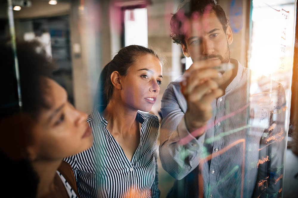 3 people stood at a glass whiteboard discussing ideas
